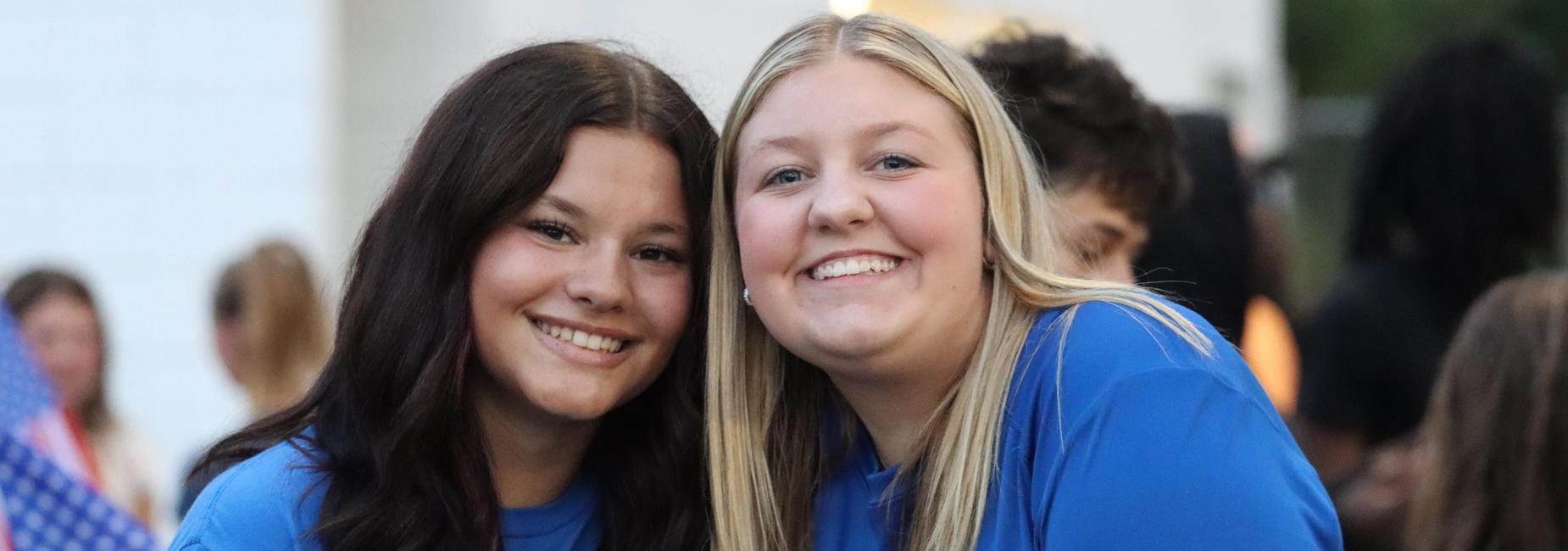 Two smiling young women in blue shirts pose together, with people in the background.