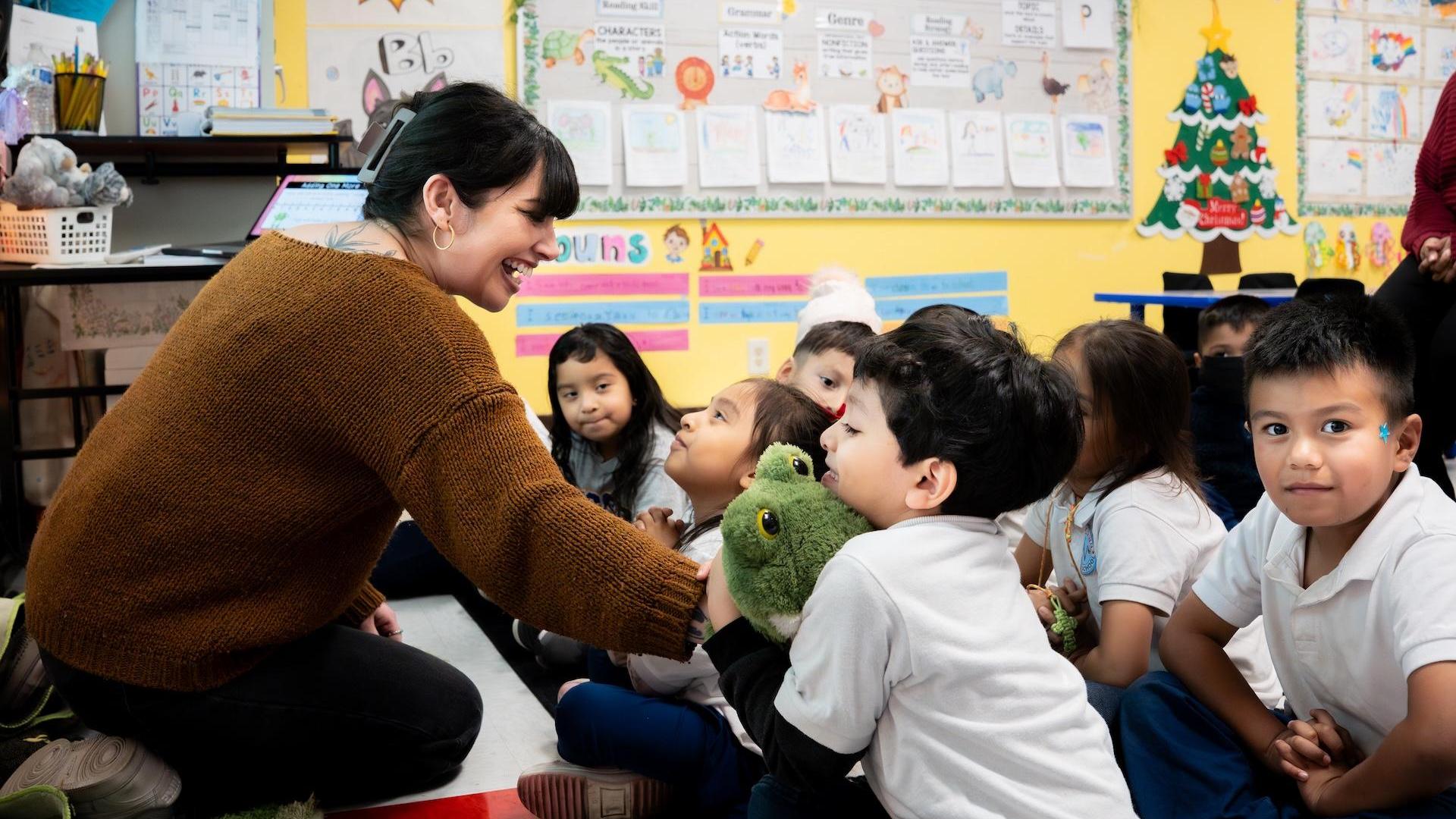 A teacher engaging with children seated on the floor in a colorful classroom.