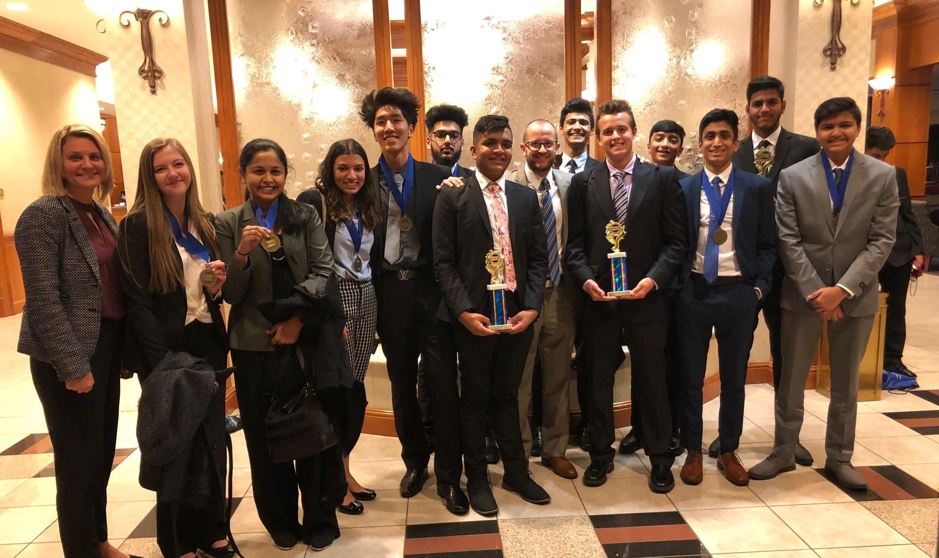 12 DECA students, 9 boys and 3 girls, holding trophies they won at the District DECA competition.