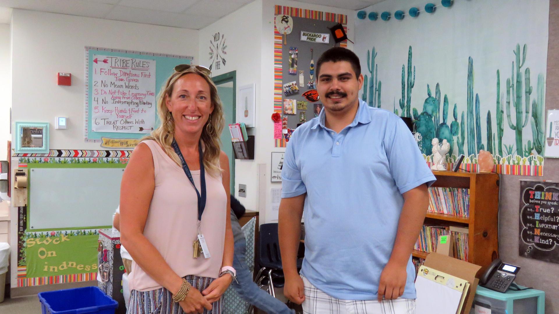 A Female and male employee smile while standing in a summer school classroom.