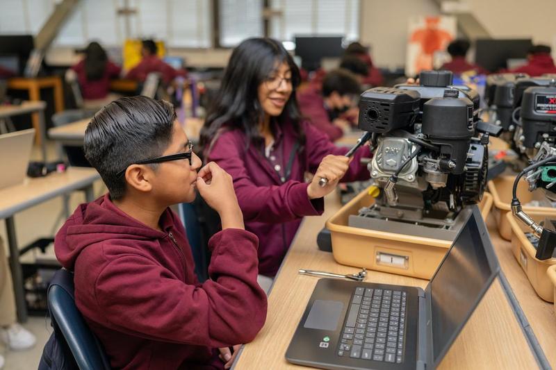 Two students at Synergy Kinetic Academy engage in a hands-on STEM activity. One student manipulates a machine while another is using a laptop. They are wearing maroon uniform shirts.