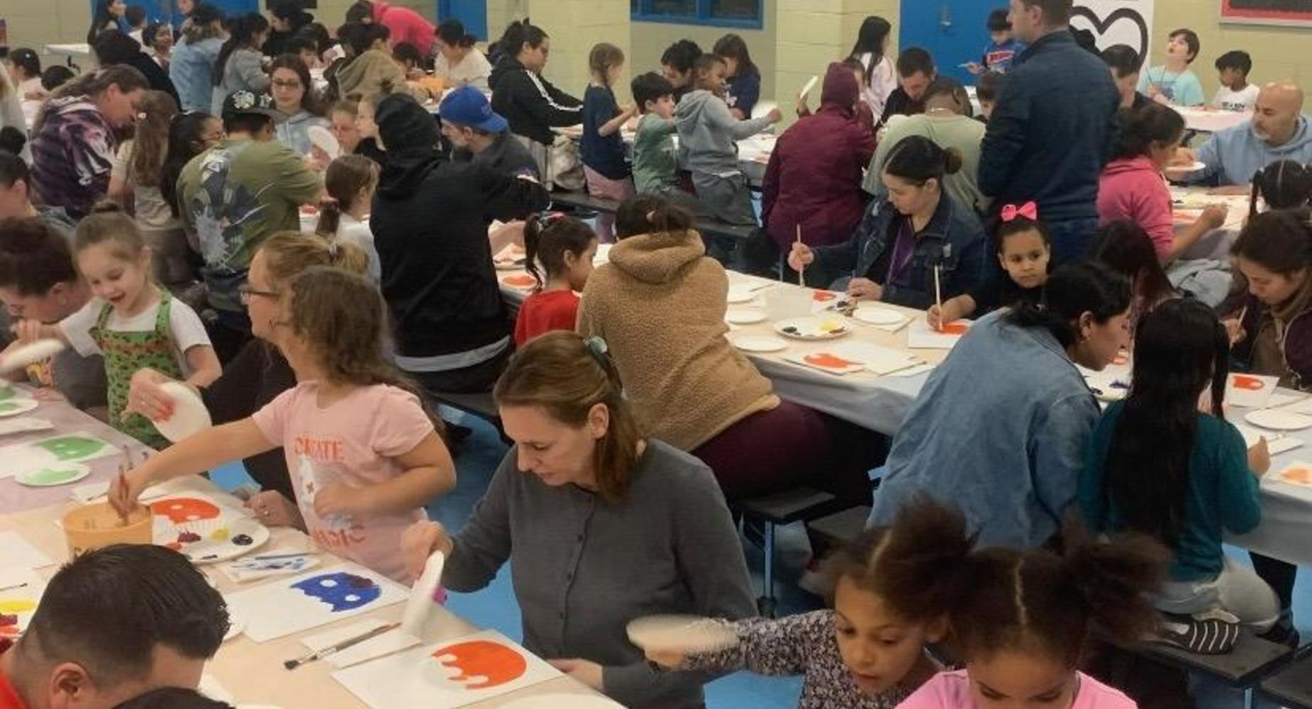 Picture of parents and students painting pictures sitting at the tables in the cafeteria