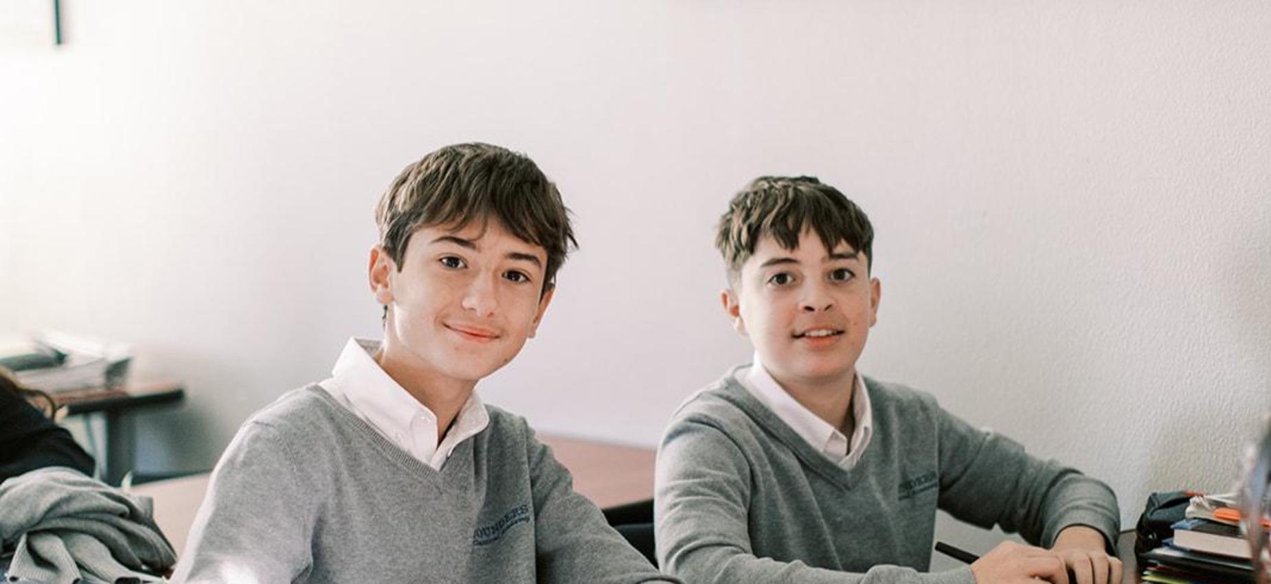 Two smiling students in gray sweaters sit at their desks