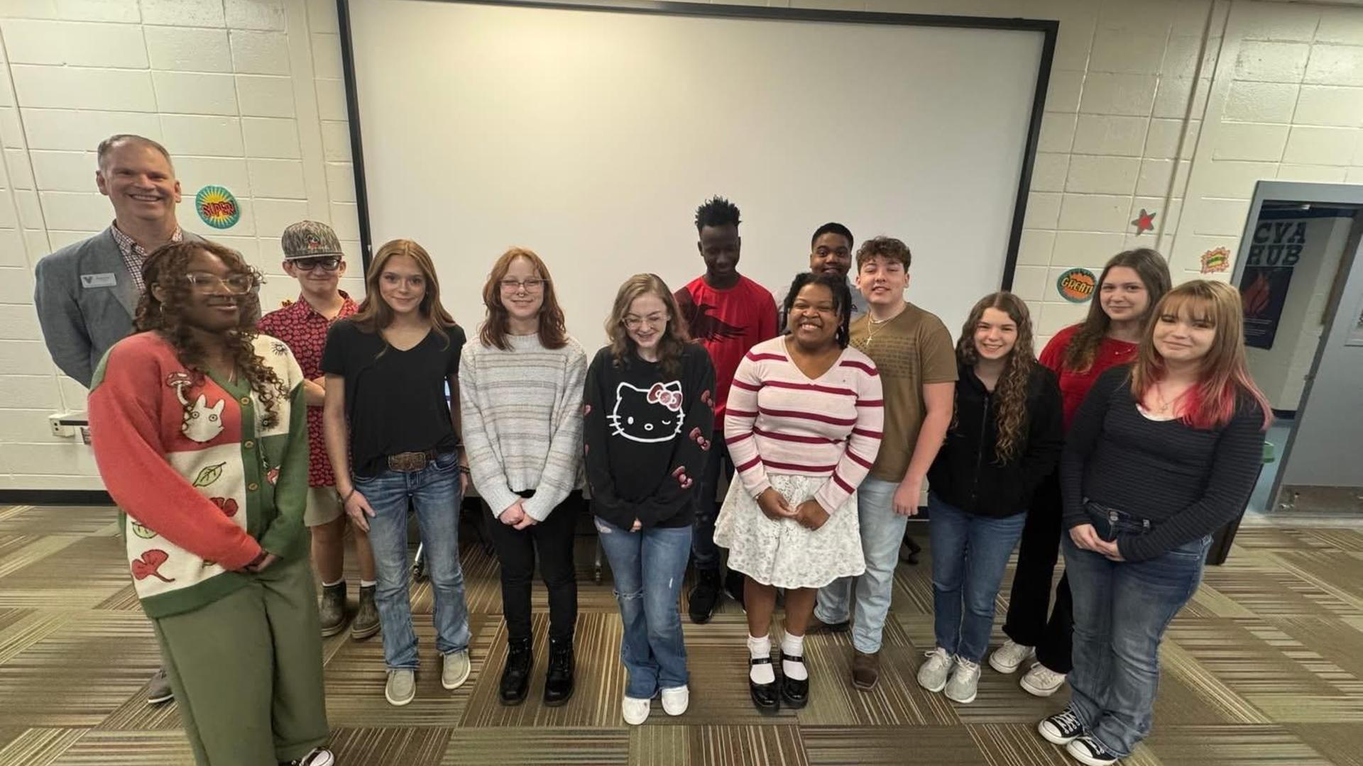 A group of diverse teenagers and adults standing together in front of a blank screen.