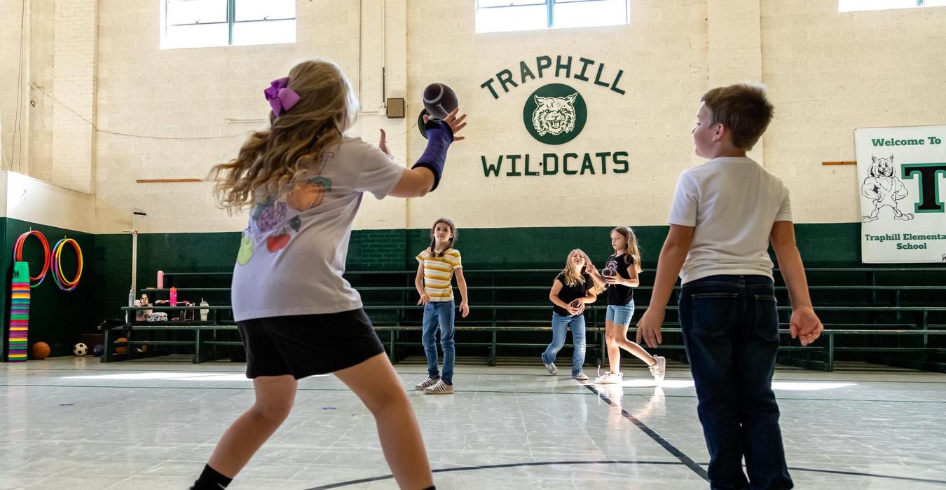 Children throwing a football in a gymnasium with a school sign behind them.