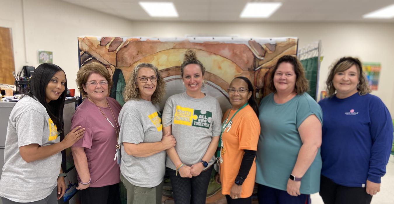 Seven women in matching shirts smiling together in front of a backdrop.