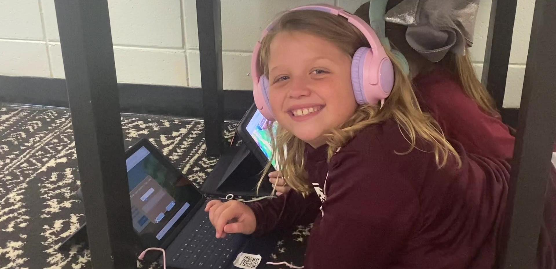Girl with pink headphones working on a tablet under a table, smiling at the camera.