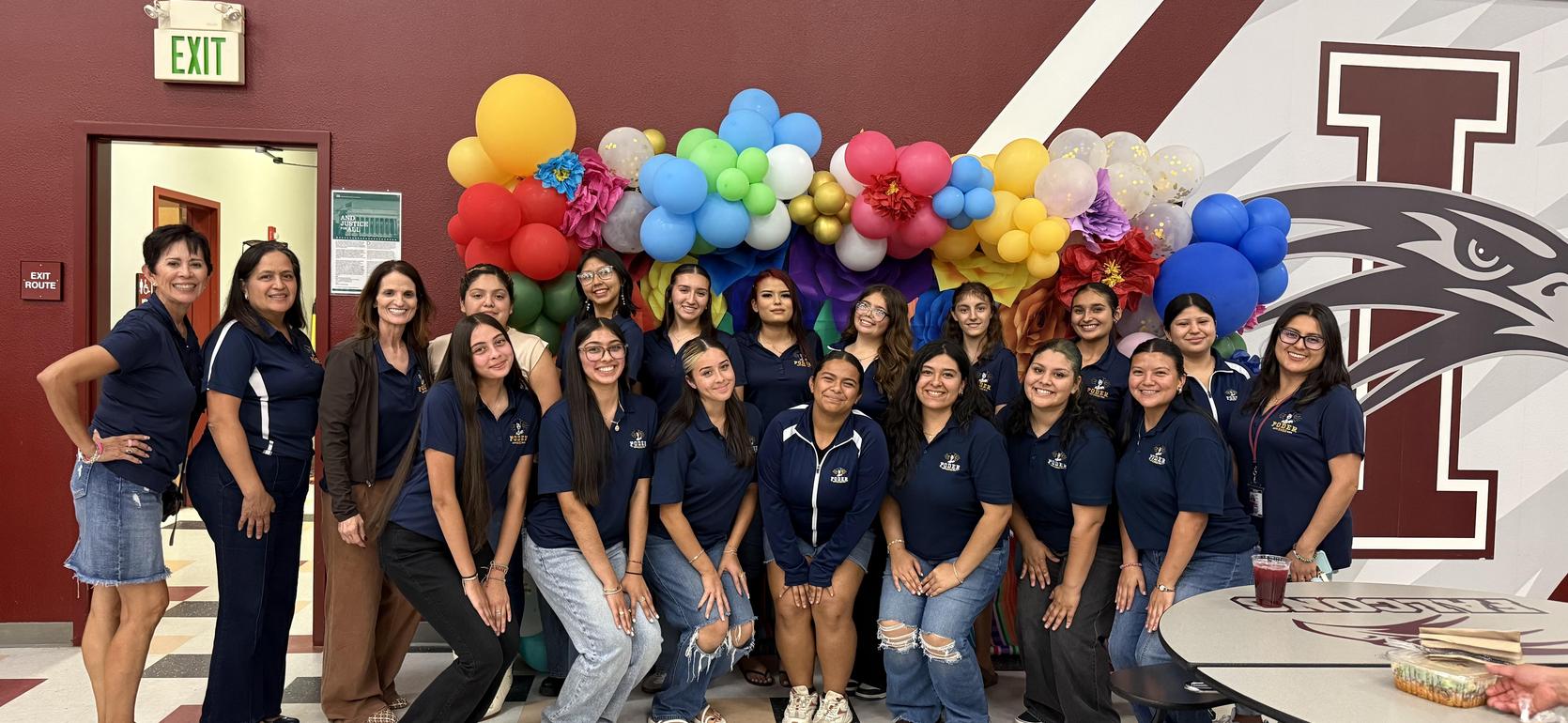 Group of students in navy shirts posing in front of a colorful balloon backdrop.