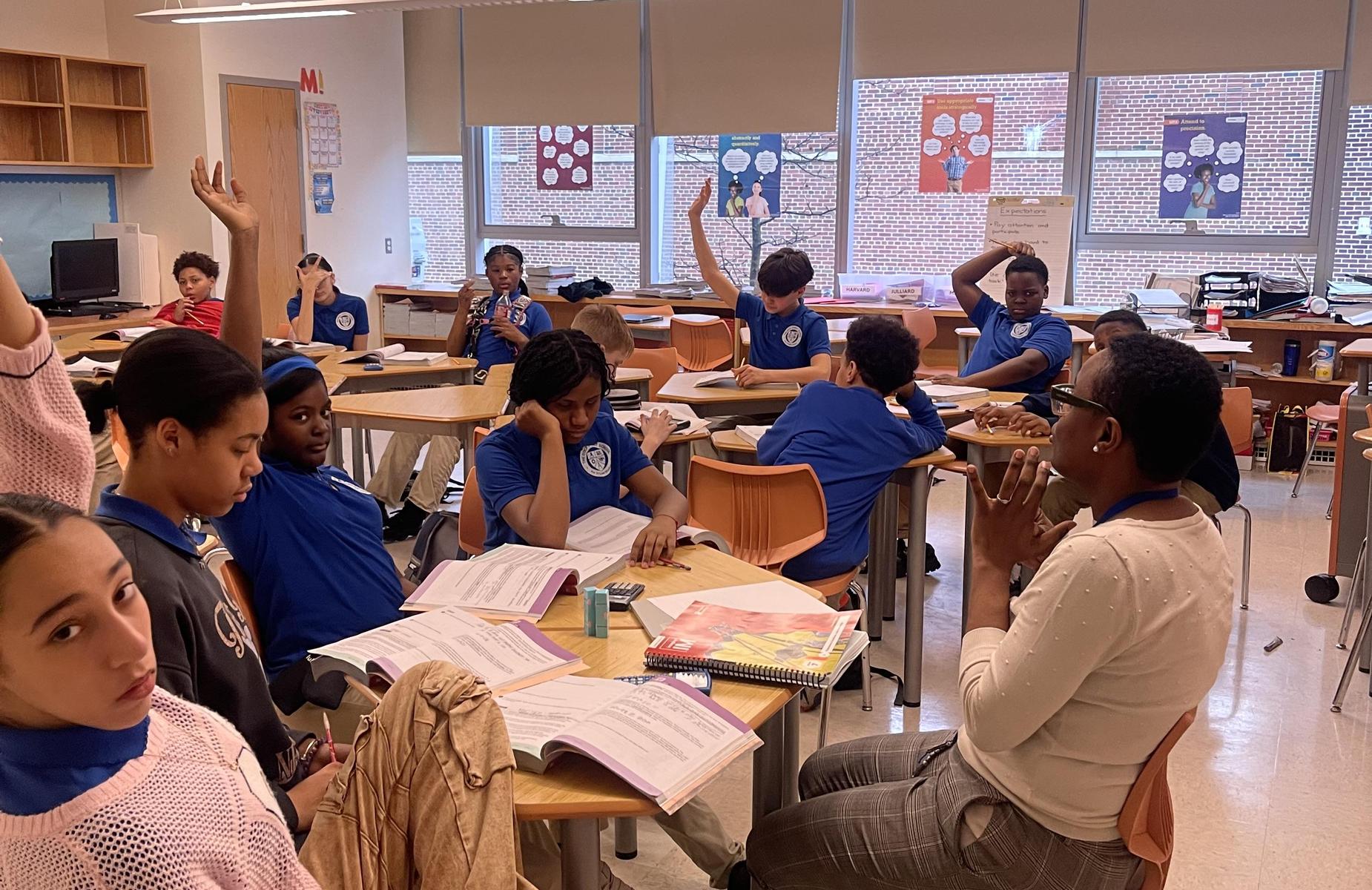 Students engaging in class, some raising hands, in a brightly lit classroom.