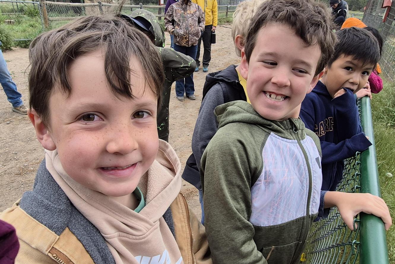 Two smiling boys in casual clothing standing by a green fence.
