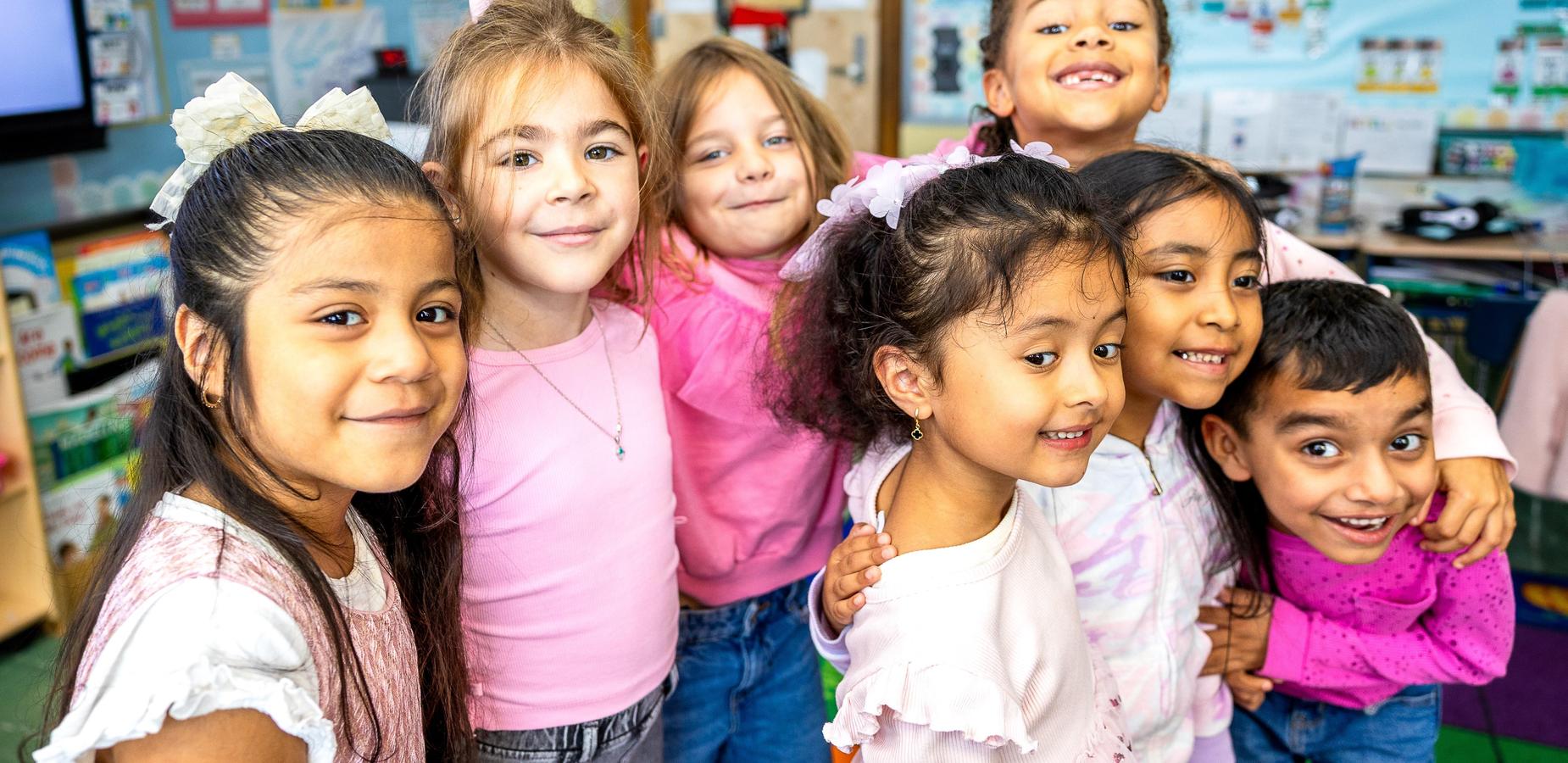 Group of seven smiling children posing together in a colorful classroom.