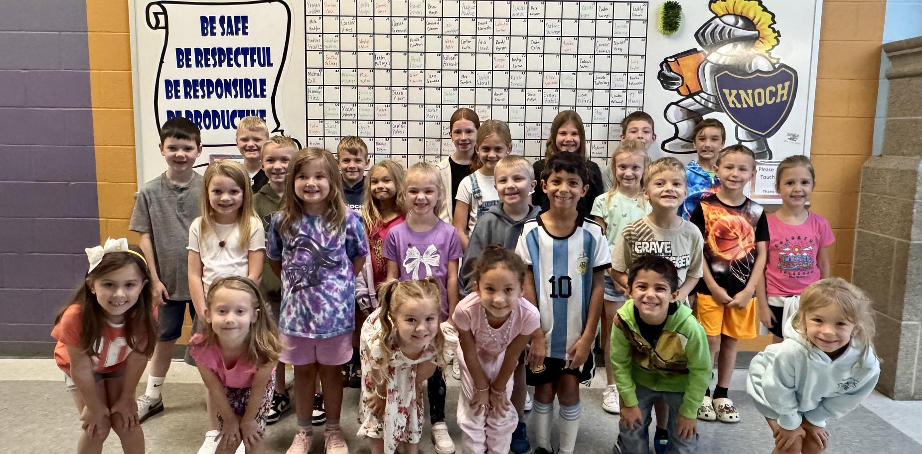 Group of smiling children posing in a school hallway with a colorful calendar.
