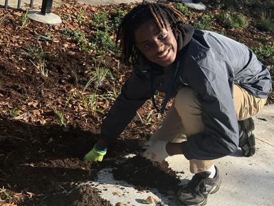 Boy planting flowers in a garden, wearing gloves and smiling at the camera.