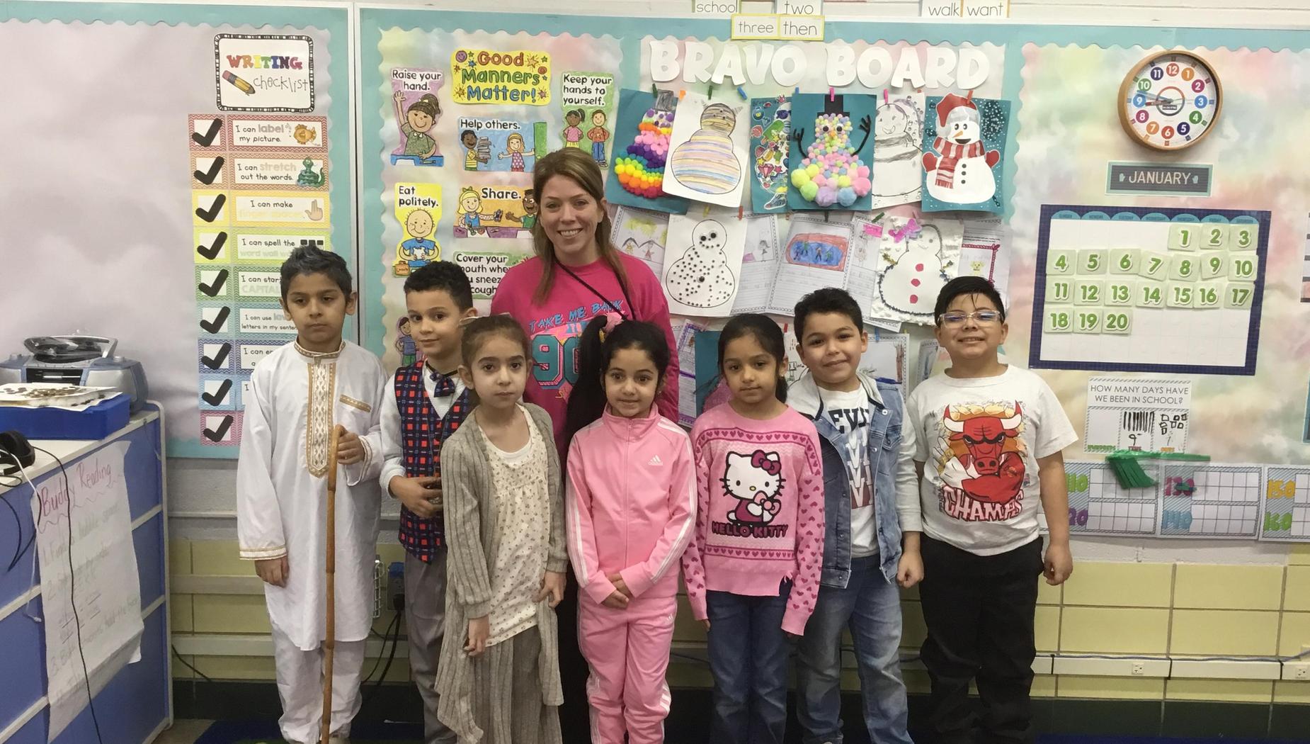 A group of children and a teacher posing in front of a colorful classroom bulletin board.
