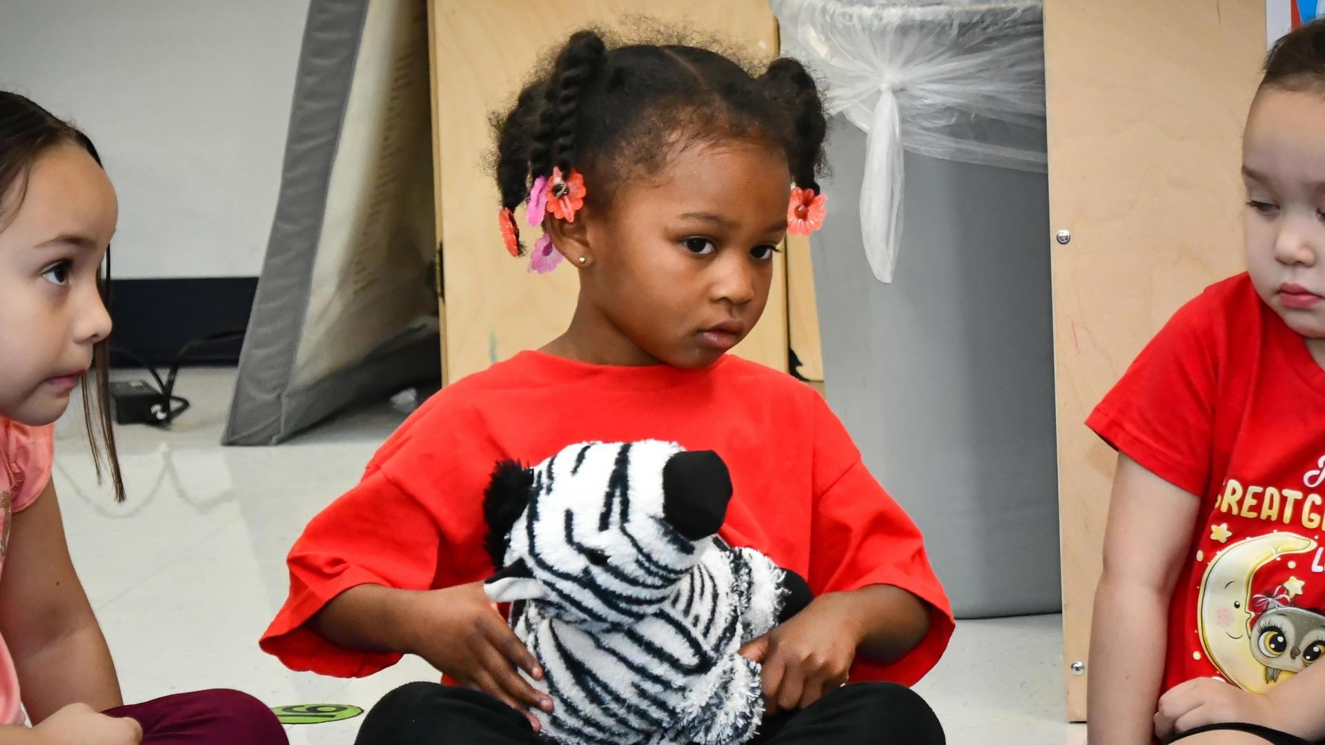 A girl in a red shirt sits with a zebra toy, looking pensively at the camera.