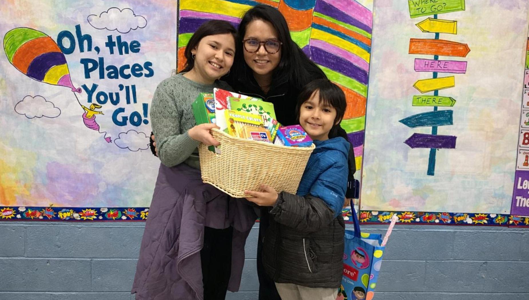 Two children and an adult hold a basket of books in front of a colorful wall.