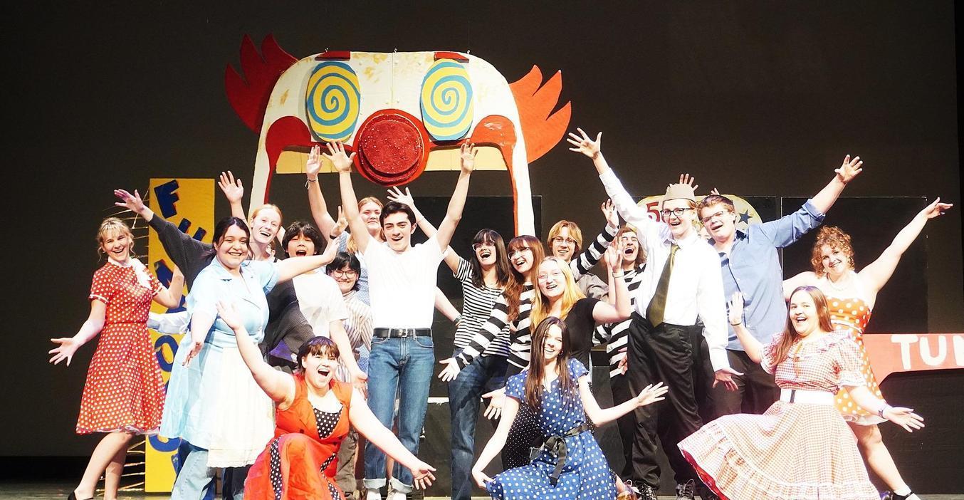 A large group of performers in colorful costumes posing cheerfully on stage.