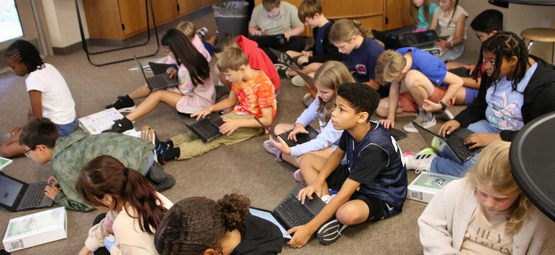 Children working on laptops while sitting on the classroom floor.