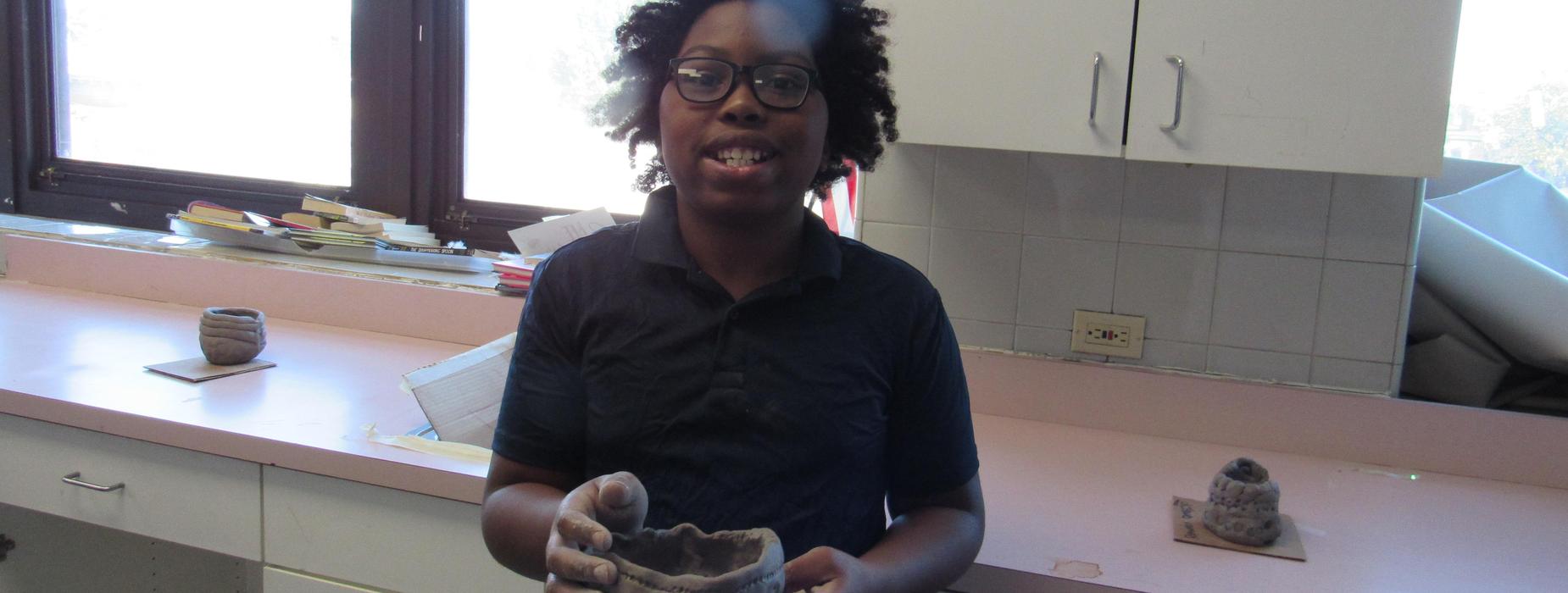 A child with curly hair holding a clay pot in a classroom setting.