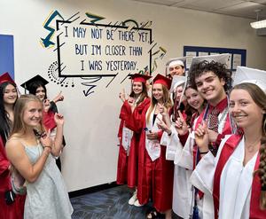 Students in graduation caps and gowns point toward an inspirational quote mural on a school hallway wall.