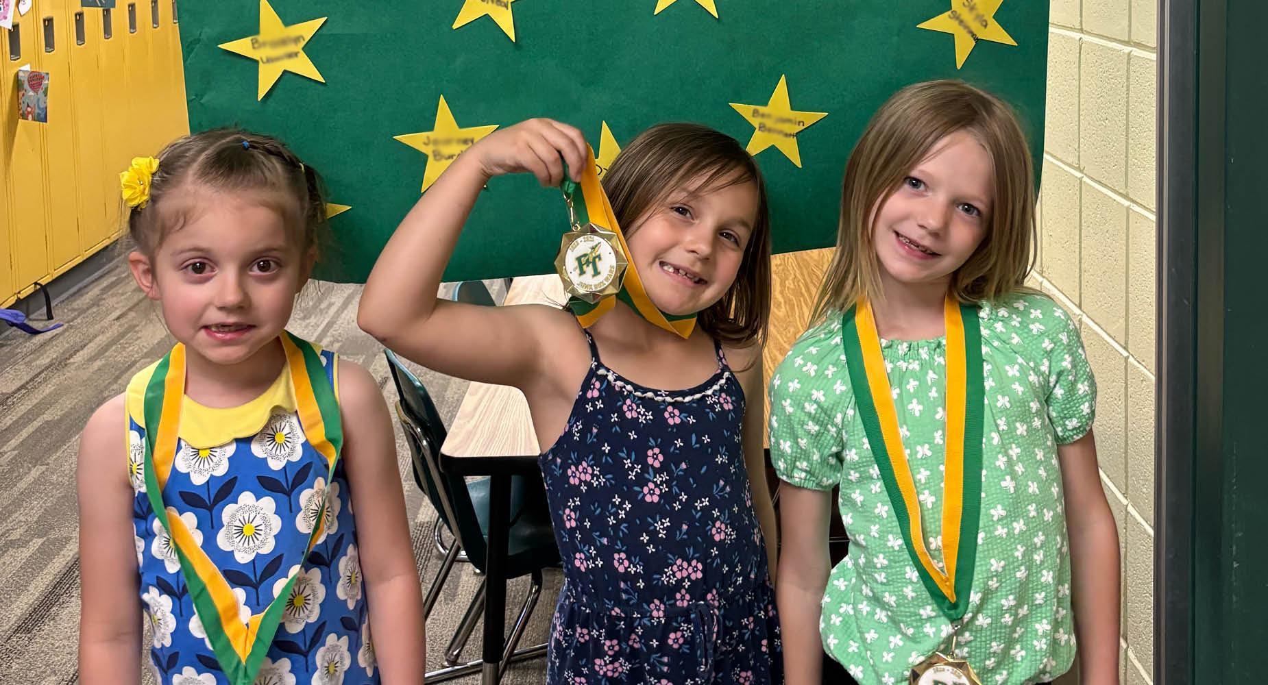 Three young girls stand in front of a green sign with yellow stars and display the medals hanging around their necks with green and yellow ribbons