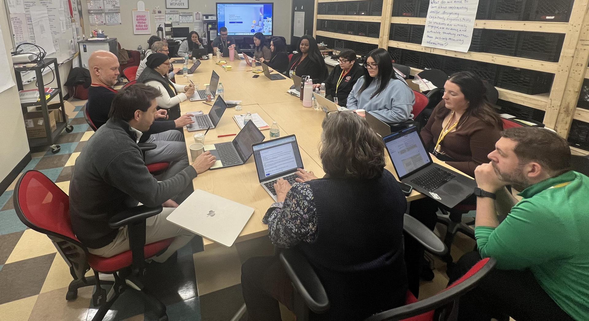 A meeting of adults seated around a long table, engaging with laptops and documents.