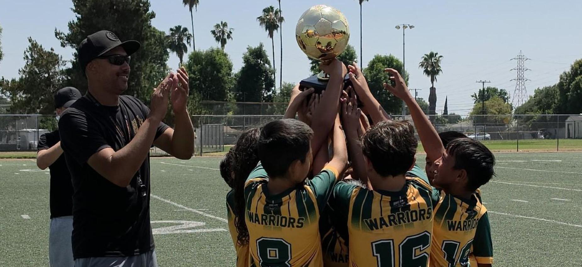 Children in soccer uniforms celebrate holding a trophy on a field.