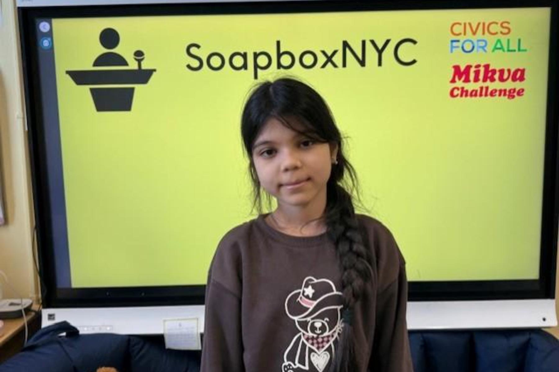 A girl in a brown shirt stands in front of a bright yellow backdrop with logos for SoapboxNYC and Mikva Challenge.