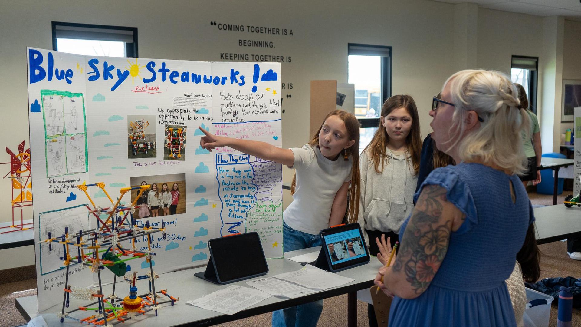 A group students presenting their project during the K&rsquo;NEX STEM Design Challenge. Students are standing behind a table displaying a colorful K&rsquo;NEX model, while one of them points to a large tri-fold presentation board that includes photos, diagrams, and handwritten notes explaining their design and the science behind it as an IU5 staff judge stands across from them, listening attentively and holding a clipboard.