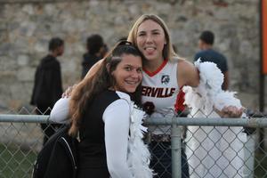 Two girls in cheerleading outfits smile and pose by a chain-link fence.