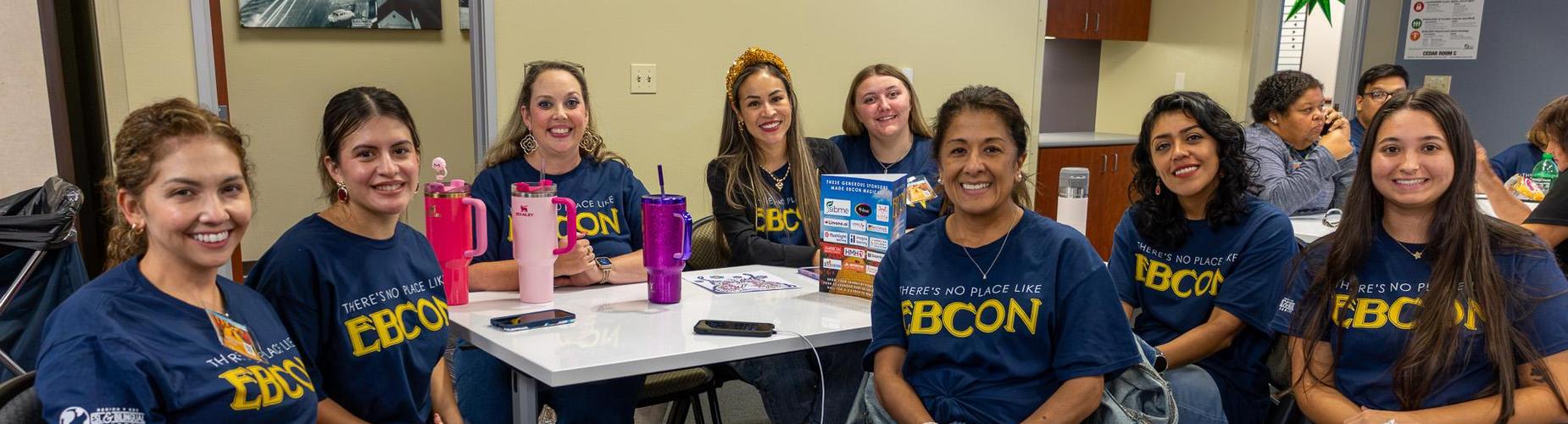 Group of smiling women in matching blue shirts, gathered around a table.