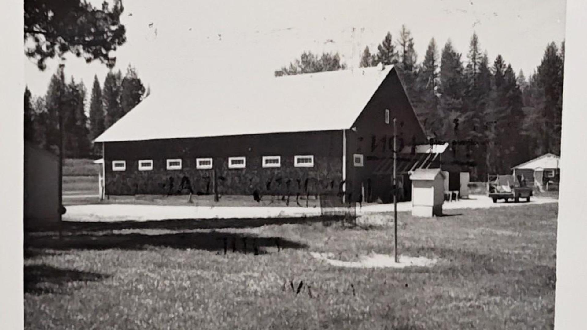 Black and white image of a red barn or large building with trees in the background.