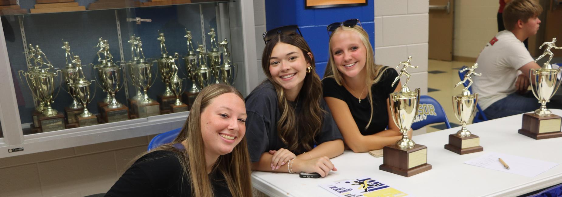Three young women smiling at a table with trophies and awards in the background.