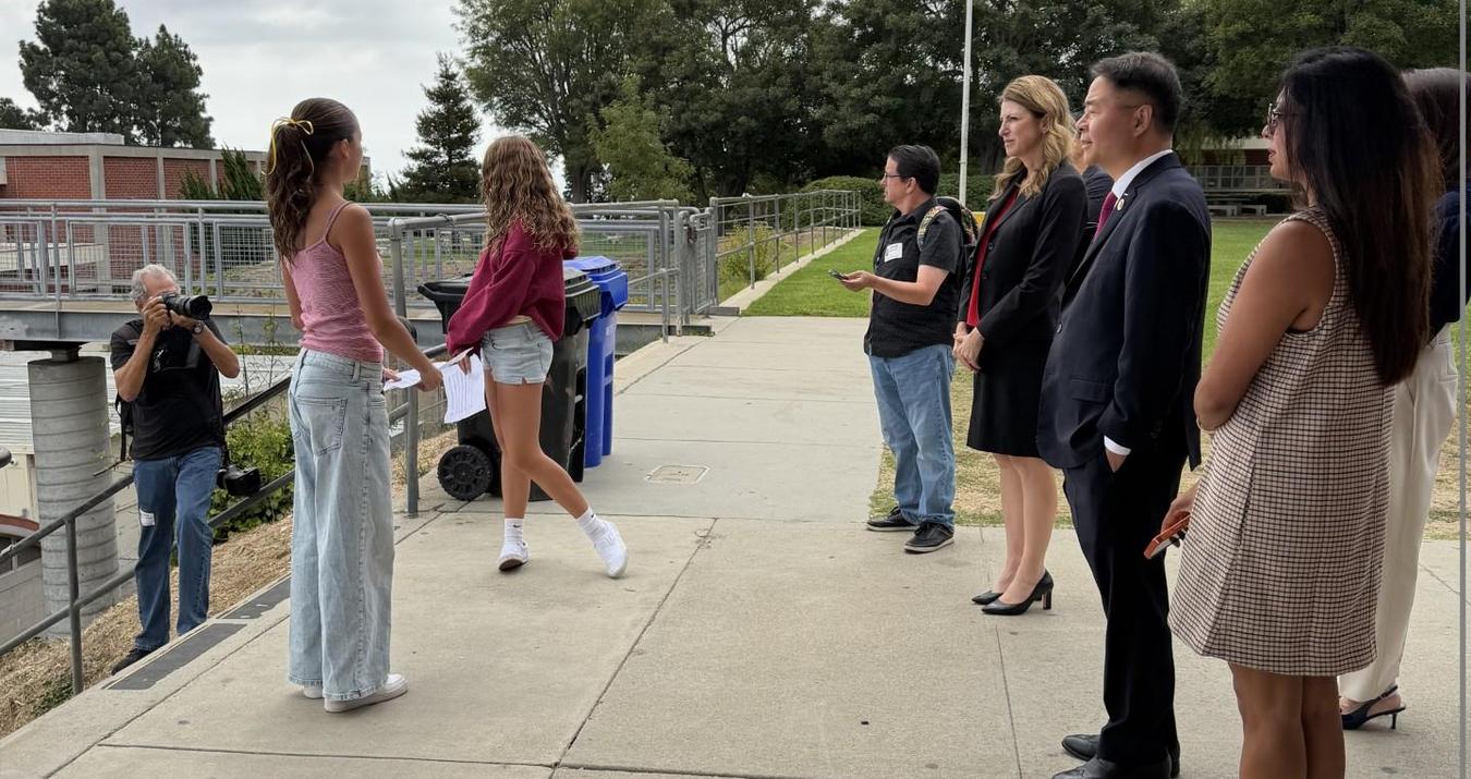Two young women interact while others observe in an outdoor setting.
