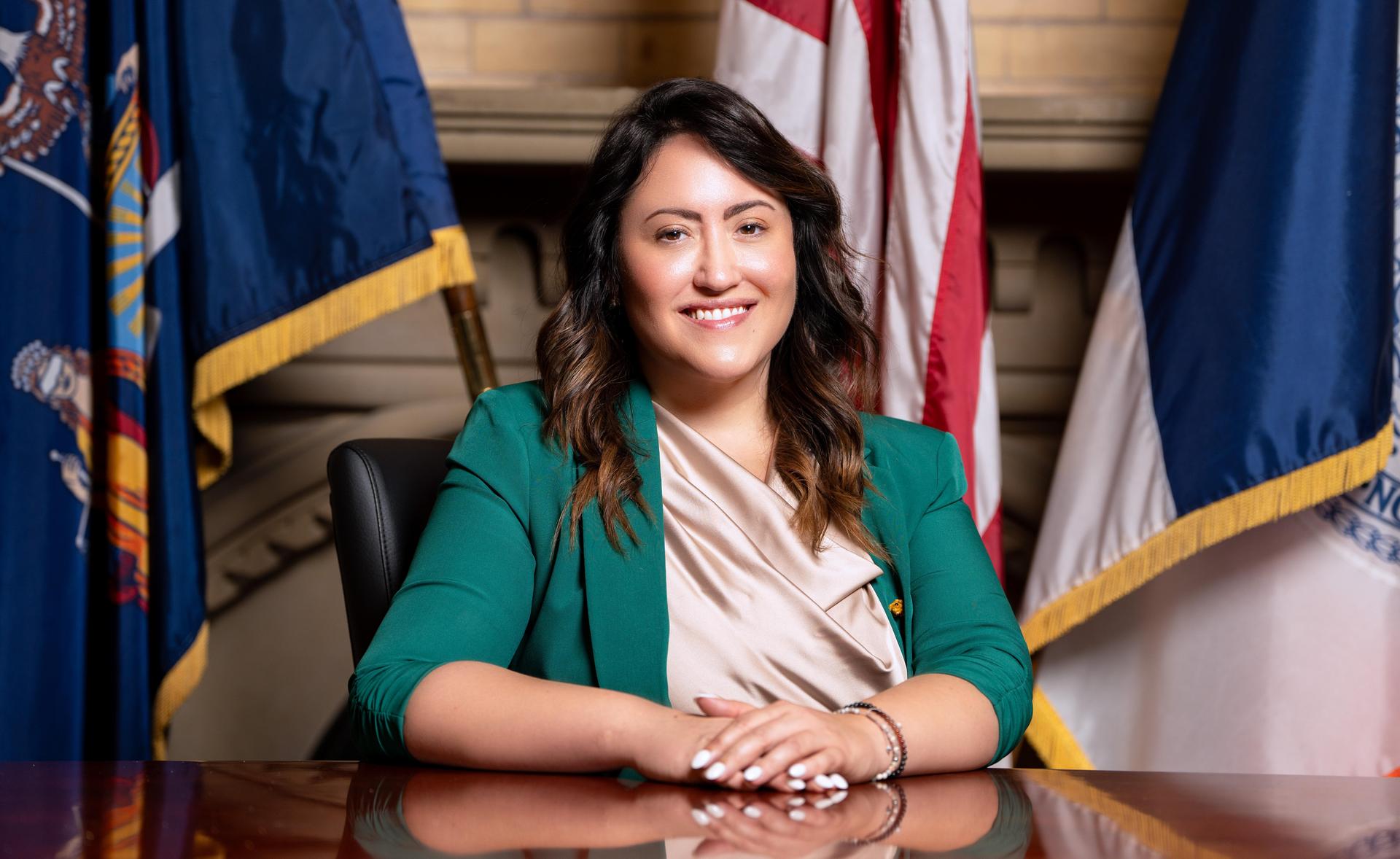 A smiling woman in a green blazer seated at a table, flags in the background.