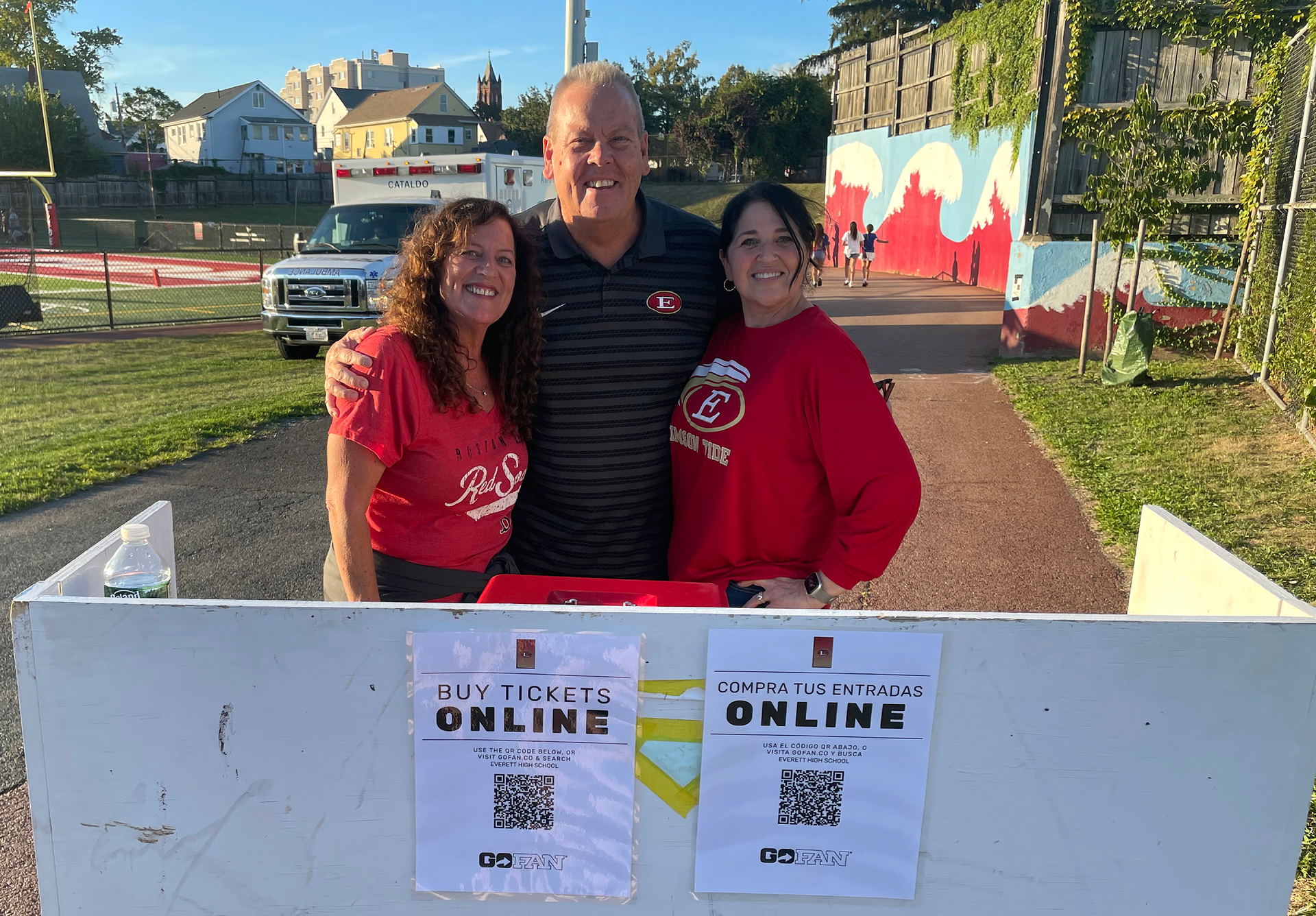 Two women and a man at an outdoor ticket booth with event signage.