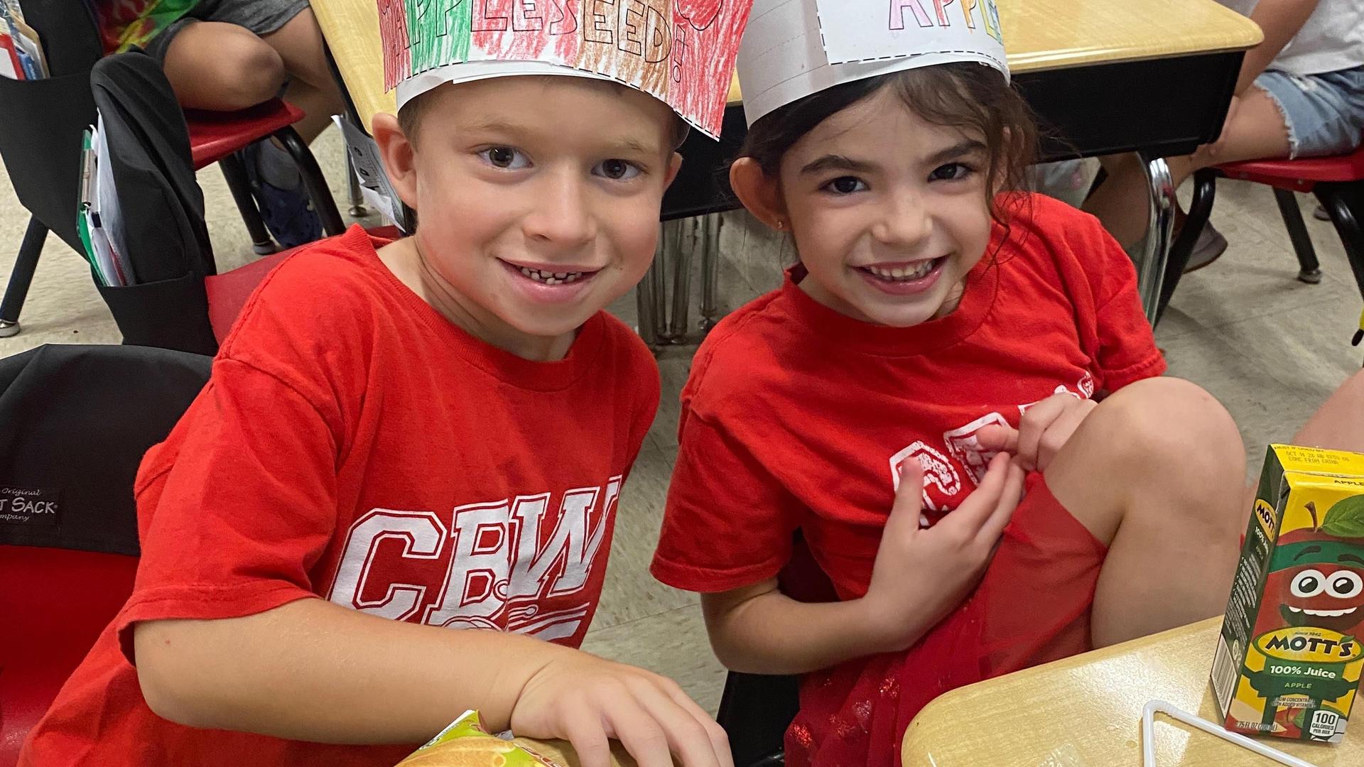 Two children wearing colorful hats and red shirts smiling at the camera in a classroom.