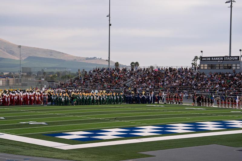 Multiple school marching bands in colorful uniforms lined up on a football field performing for a packed crowd.