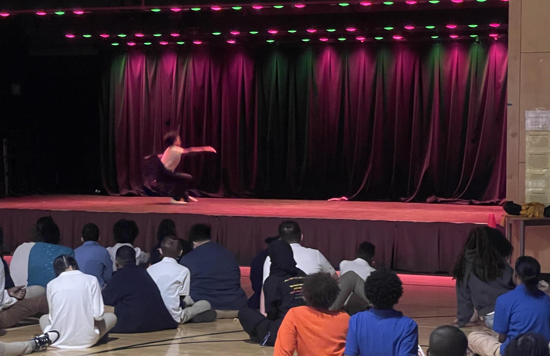 A dancer performs on stage with colorful lighting, while an audience watches.