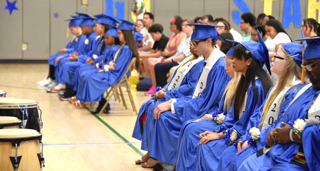 Graduating students in blue caps and gowns seated in a gym, some focused ahead.