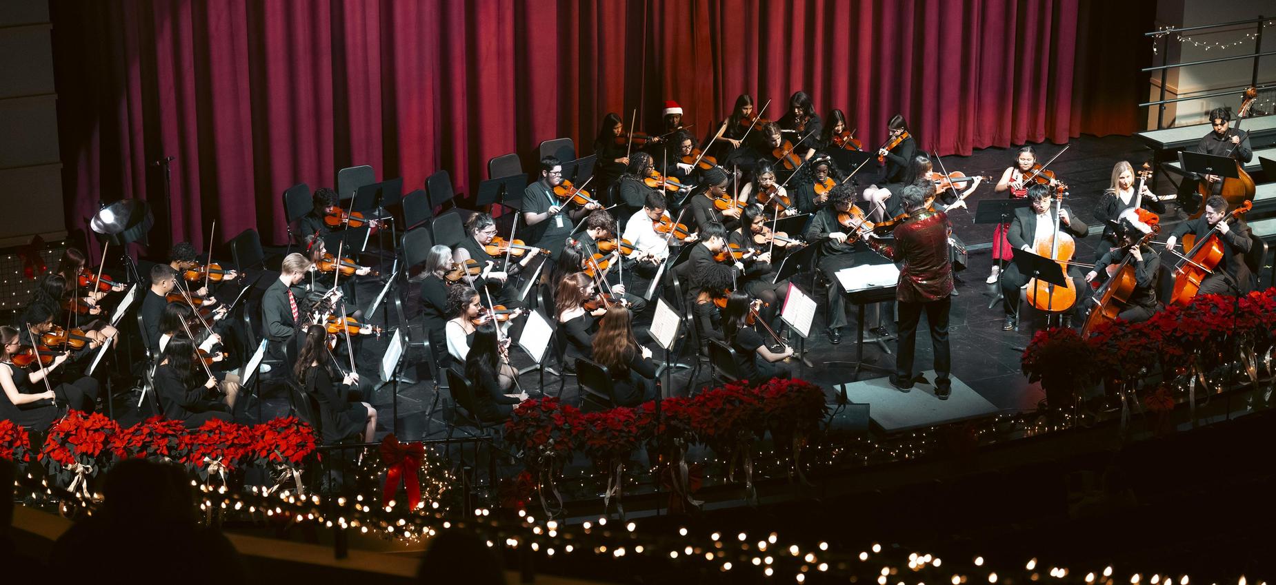 Orchestra performing on stage with musicians and a conductor, decorated with garlands.