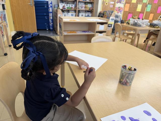 Child drawing at a classroom table with colorful supplies around.