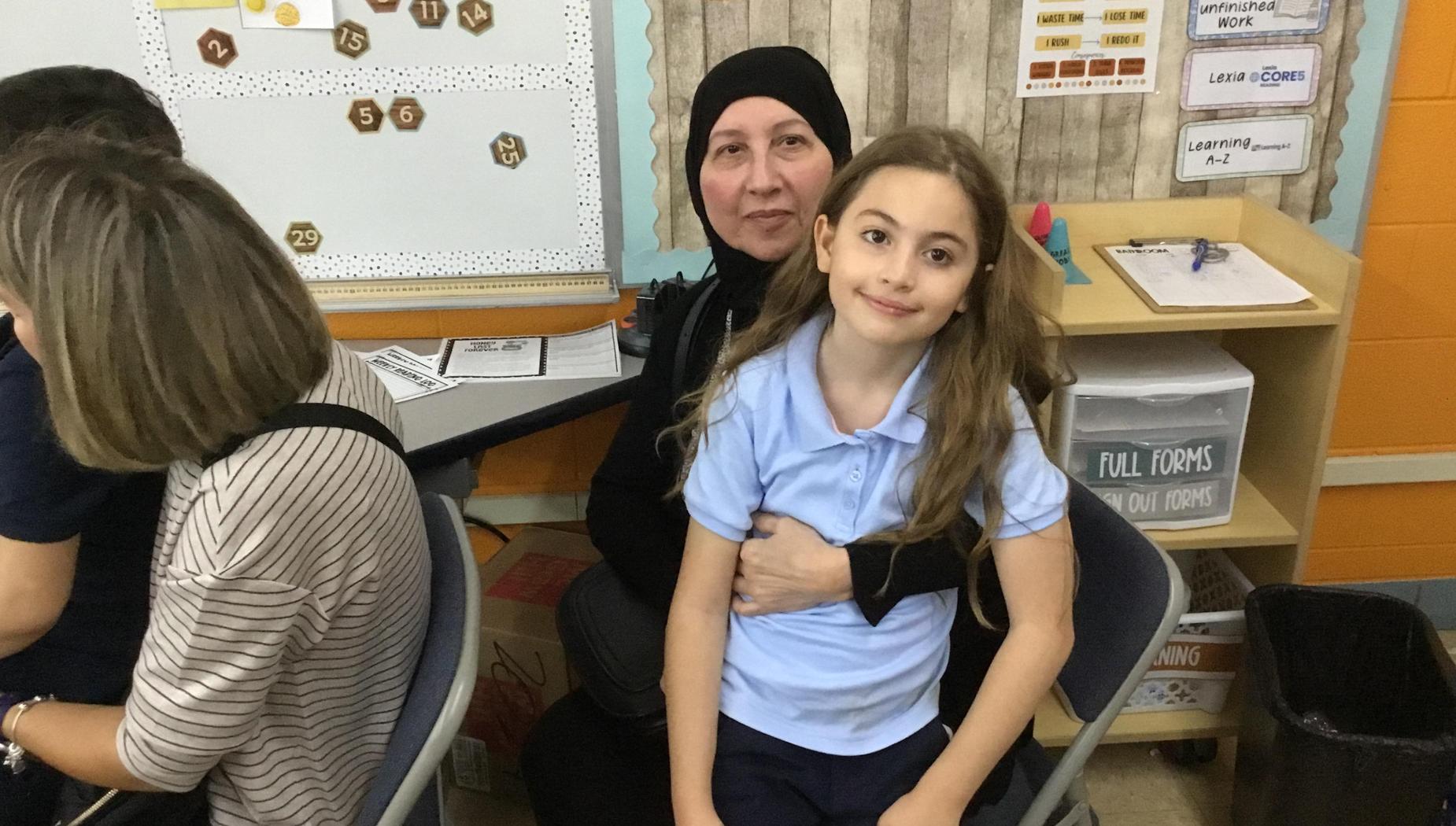 A woman in a black hijab poses with a girl in a blue shirt in a classroom setting.