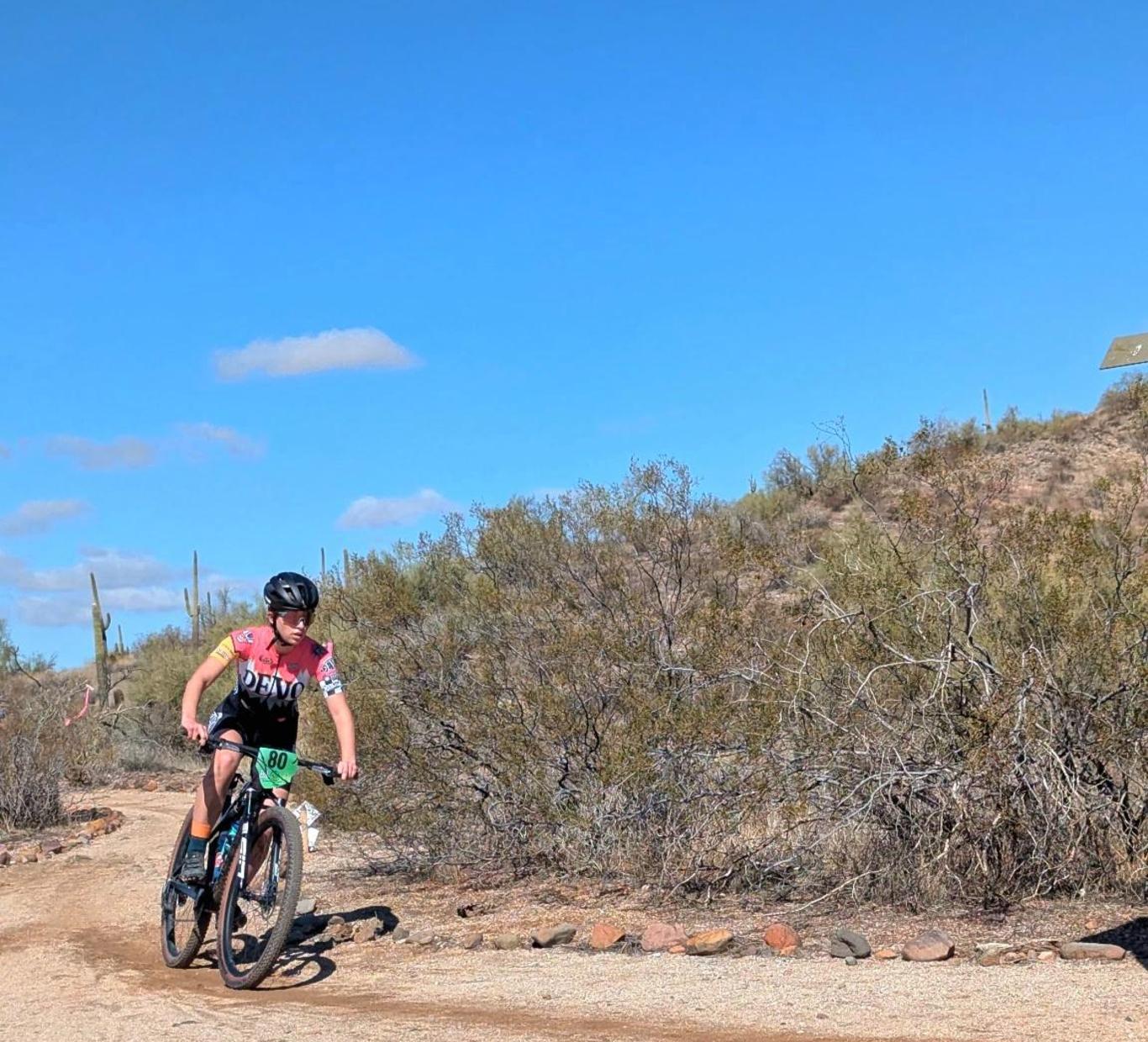 A cyclist in action on a dirt path surrounded by desert vegetation under a blue sky.