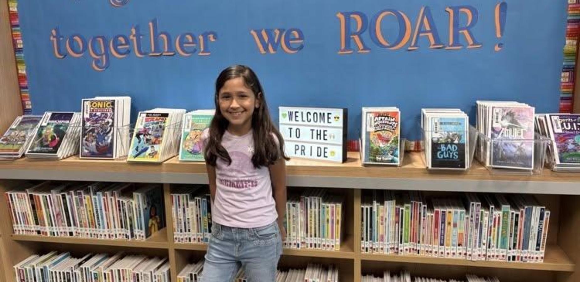 A smiling girl stands in front of bookshelves with a 'Welcome to the Pride' sign.