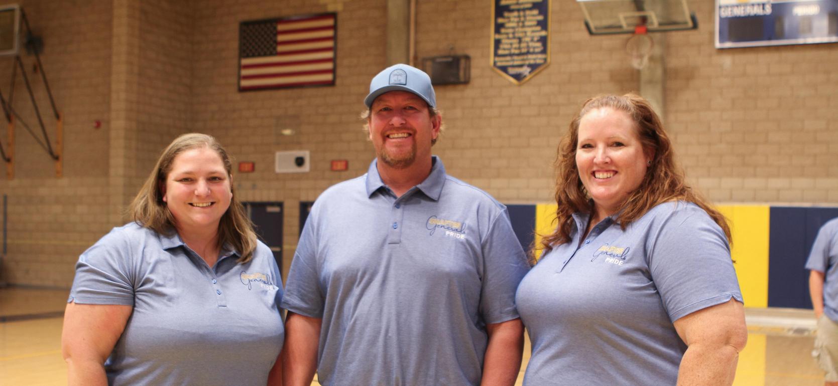 Three staff members smile together in blue shirts in a gymnasium setting.
