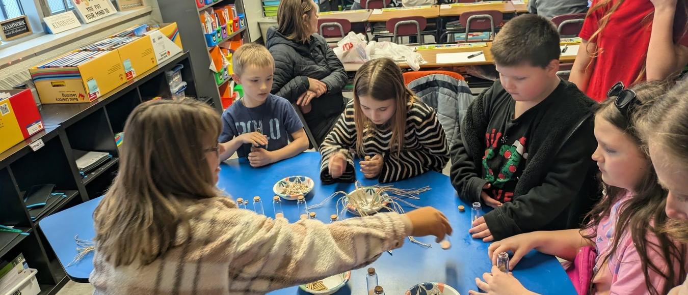 Students gather around a blue classroom table for a hands-on craft activity with jars and string.