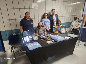 Five people stand behind a table with promotional materials and a game wheel.