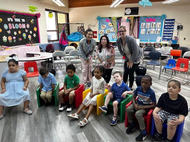 a group of preschool age children sit in a row of chairs in front of their teachers who are standing behind them