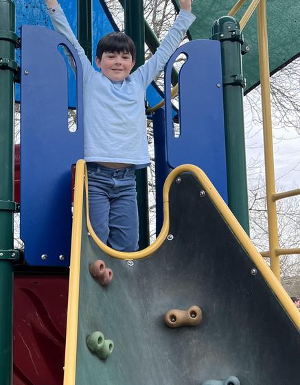 Child standing on a slide at a playground, raising arms in excitement.